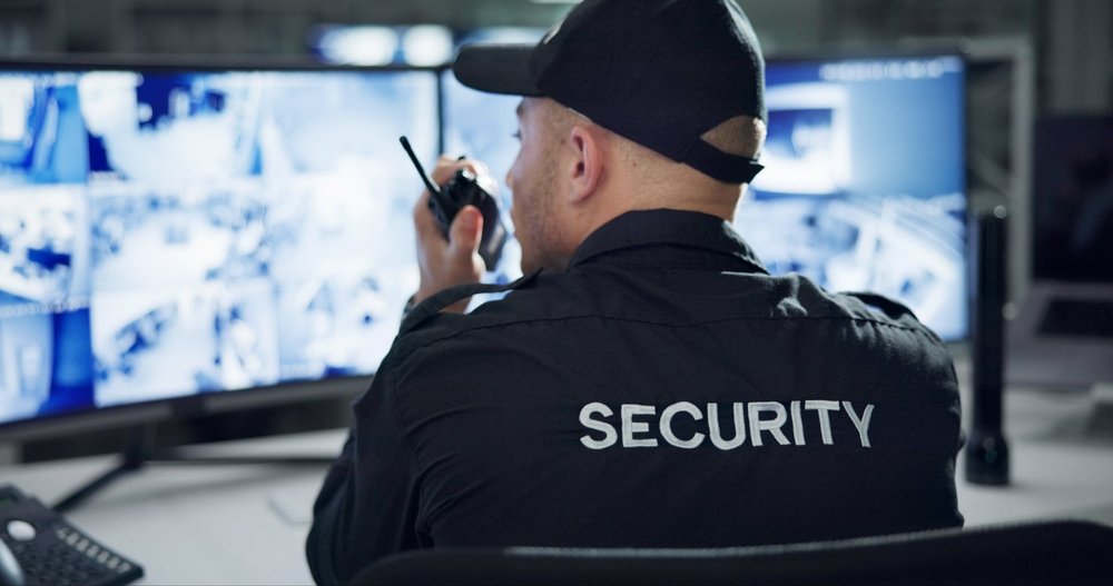 A security officer, an integral part of business security, wears a uniform with "SECURITY" emblazoned on the back and holds a walkie-talkie while diligently monitoring multiple surveillance screens displaying various company locations.