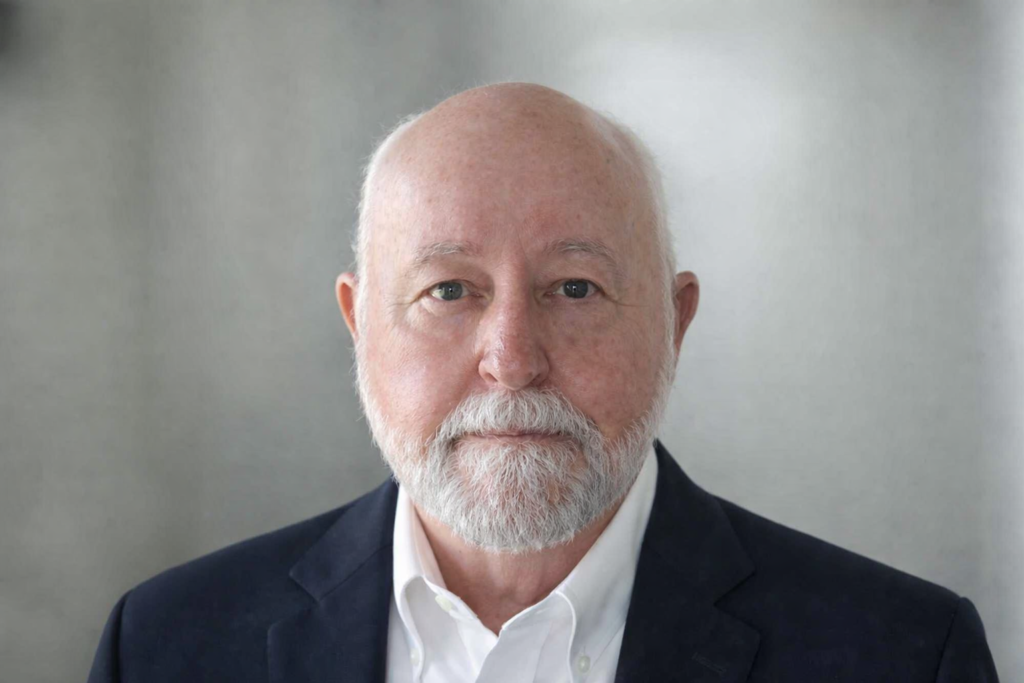An older man with a bald head and white beard, wearing a dark suit jacket and white shirt, looks directly at the camera against a neutral, blurred background.